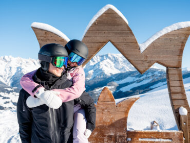 Twee mensen in ski-uitrusting glimlachen op een besneeuwde berg met houten decor, genietend van een heldere winterdag.