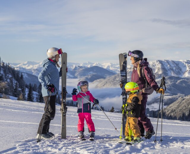 Twee volwassenen en twee kinderen in skikleding staan op een besneeuwde berg met ski’s en skistokken, met bergen op de achtergrond.