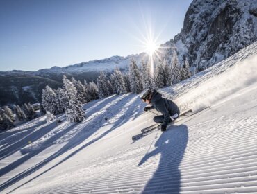 Een skiër die over een geprepareerde besneeuwde helling carvet, terwijl de zon schijnt over de besneeuwde bomen en bergen.