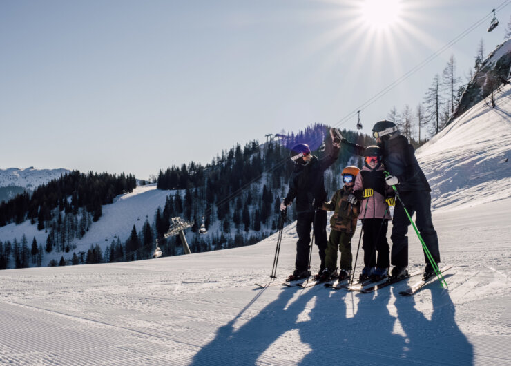 Vier mensen in ski-uitrusting staan op een besneeuwde helling met bomen en bergen op de achtergrond onder de felle zon.