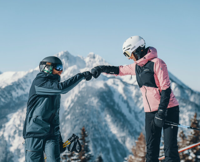 Twee skiërs in winteruitrusting maken een vuist op een besneeuwde berg met schilderachtige bergtoppen op de achtergrond.