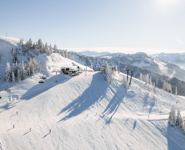 Besneeuwde bergpiste met skiërs, skiliften en een lodge onder een heldere hemel met uitzicht op de bergen.