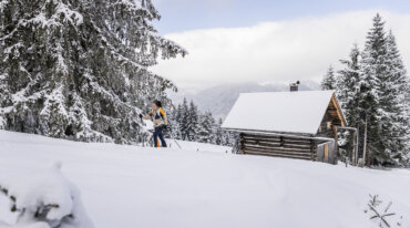 Iemand skiet bergopwaarts langs een met sneeuw bedekte hut en dennenbomen in een winters berglandschap.