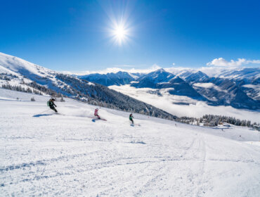 Drie skiërs dalen een besneeuwde berghelling af onder een stralende zon met bergen en wolken op de achtergrond.