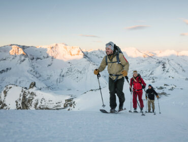 Drie mensen in winteruitrusting op sneeuwschoenen op een besneeuwde berghelling met besneeuwde bergtoppen op de achtergrond.