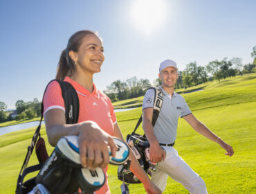 Golfen in het SalzburgerLand Twee golfers lachen en lopen op een zonnige golfbaan, elk met een golftas over hun schouder.