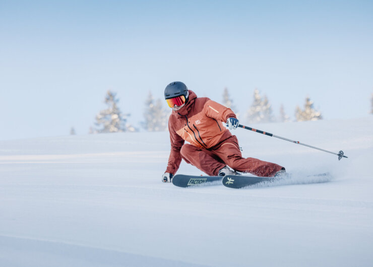 Skier in een oranje ski-outfit maken een soepele carve-bocht op een goed geprepareerde piste bij mooi winterweer.