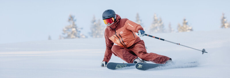 Skier in een oranje ski-outfit maken een soepele carve-bocht op een goed geprepareerde piste bij mooi winterweer.