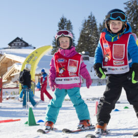 Twee blije kinderen tijdens de kinderskicursus met skikleding, ski’s en helm. Op de achtergrond is een funpark voor kinderen te zien.