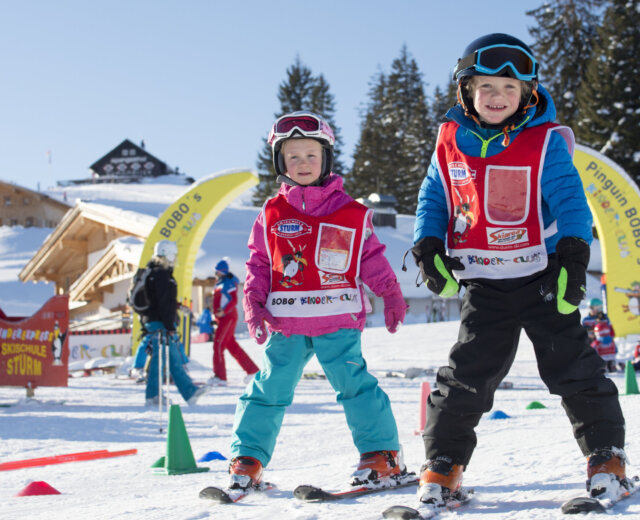 Twee blije kinderen tijdens de kinderskicursus met skikleding, ski’s en helm. Op de achtergrond is een funpark voor kinderen te zien.