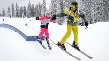 Een kind wordt door papa aan de hand gehouden terwijl het lachend en goedgemutst in een kinderpark met ski’s over een hindernis glijdt.