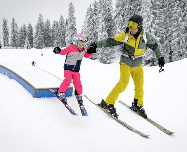 Spaß beim Skifahren im Kinderpark-Dachstein_West_(c)_Mirja_Geh Een kind wordt door papa aan de hand gehouden terwijl het lachend en goedgemutst in een kinderpark met ski’s over een hindernis glijdt.