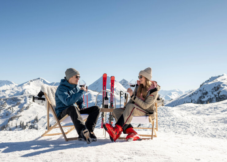 Een man en een vrouw zitten aan een klein tafeltje op de skipiste, met hun ski’s achter hen geparkeerd, en genieten van een glas wijn bij prachtig weer.