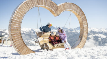 Een vader, een moeder en hun twee kinderen nemen een pauze van het skiën, zittend in een houten schommelstoel naast een bergstation.