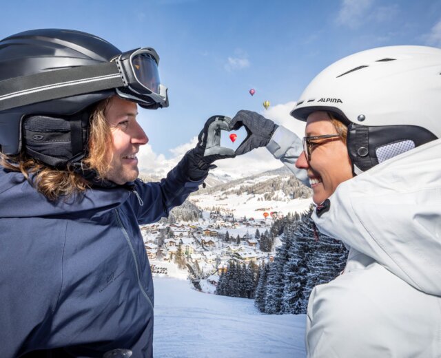 ChatGPT: Een opname van een skiër en een skiester van achteren, die elkaars hand vasthouden en samen een hart vormen – in de lucht zijn ballonnen te zien.