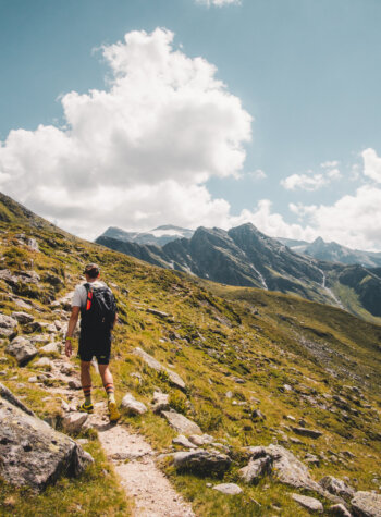 Een persoon wandelt over een rotsachtig bergpad onder een heldere hemel met wolken en verre bergtoppen.