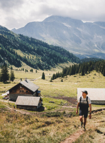 Een persoon wandelt op een met gras begroeide heuvel in de buurt van berghutten, omringd door bomen en hoge bergtoppen onder een bewolkte hemel.
