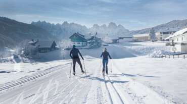 Twee langlaufers op de loipe in Gosau – de één in klassieke stijl, de ander skatend – genieten van het uitzicht op besneeuwde bergtoppen onder een stralende zon.