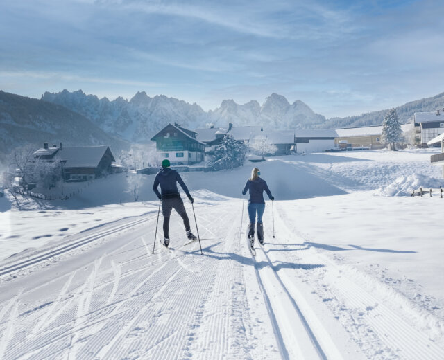 Langlaufen_Gosau (c)Dachstein West Twee langlaufers op de loipe in Gosau – de één in klassieke stijl, de ander skatend – genieten van het uitzicht op besneeuwde bergtoppen onder een stralende zon.