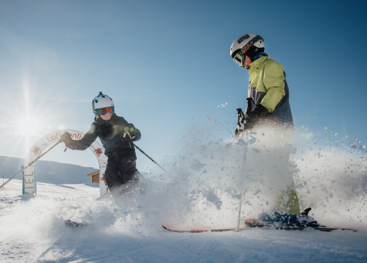 Almenwelt Lofer Lofi Funline: Een kind maakt een bocht met de ski’s, remt af en stuift sneeuw op. De vader is omgeven door een wolk van sneeuwstof.