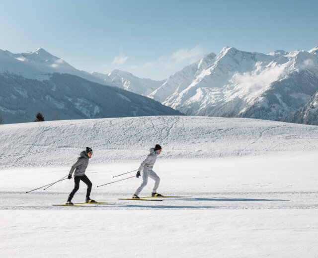 NPHT_Langlaufen am Wasenmoos oberhalb Mittersill mit Blick auf die Hohen Tauern (c) Ferienregion Nationalpark Hohe Tauern – Mathäus Gartner Twee langlaufers aan het skaten op de loipe op het Wasenmoos boven Mittersill, met uitzicht op de Hohe Tauern.