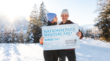 Twee personen staan midden in een met sneeuw bedekt winterlandschap en houden de Nationalpark WinterCard in de camera.