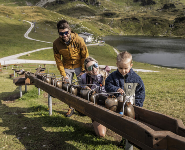 Een familie speelt met koebellen op een houten constructie in een grasveld bij een meer en bergen.