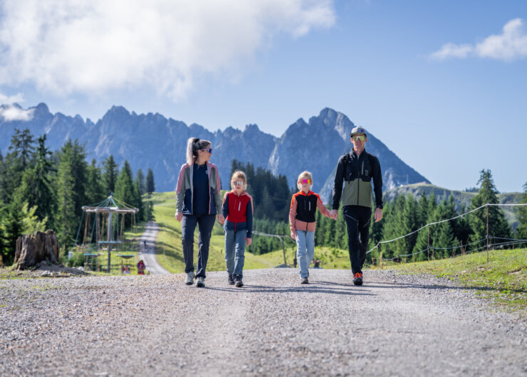 Een gezin van vier wandelt op een zonnige dag over een grindpad in de bergen, omringd door bomen en bergtoppen.