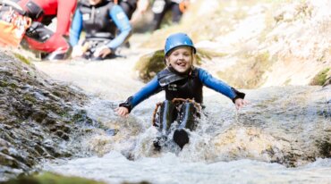 Lachend kind met helm en wetsuit glijdt langs een rotsachtige stroom met anderen op de achtergrond, genietend van canyoning.