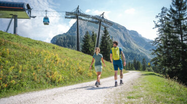 Twee mensen wandelen op een zonnige dag over een grindpad in de bergen in de buurt van een kabelbaan.