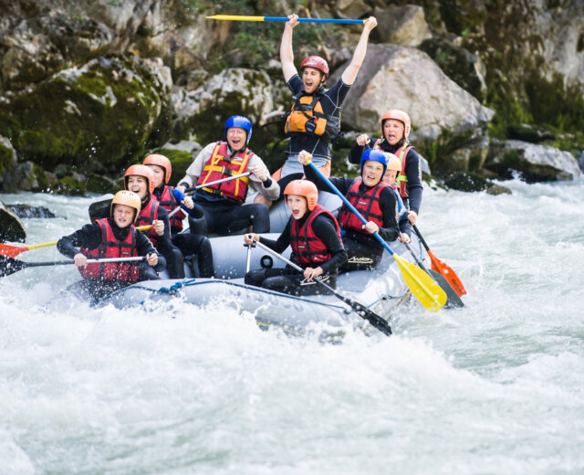 Een groep mensen met helmen op die aan het wildwaterraften zijn, lachend en juichend terwijl ze door stroomversnellingen razen.
