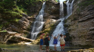 Vier kinderen met rugzakken staan bij een rotsachtige waterval en poel en kijken omhoog naar het klaterende water.