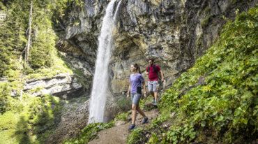 Twee mensen wandelen op een pad naast een hoge waterval en weelderige groene vegetatie in een bebost gebied.