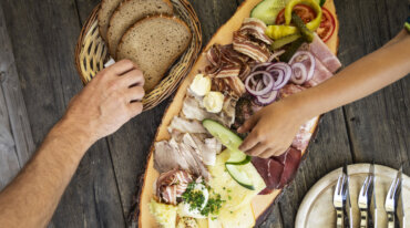 Twee handen reiken naar een houten schaal met verschillende soorten vlees, kaas, groenten en brood op een rustieke tafel.