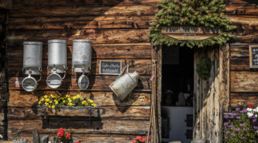 Houten gebouw met melkbussen, gele en rode bloemen, een groene krans en een open deur.