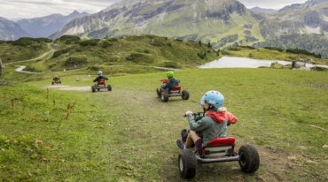 Kinderen met helmen rijden met skelters van een met gras begroeide heuvel af met bergen en een meer op de achtergrond.