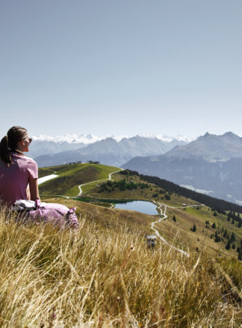 Twee mensen zitten in hoog gras op een berg en kijken uit over een schilderachtige vallei met meren, bomen en bergtoppen in de verte.