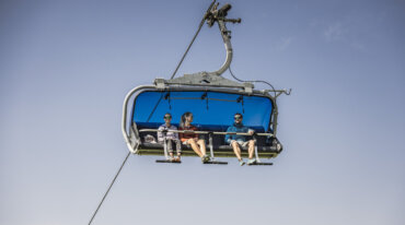 Drie mensen rijden op een blauwe stoeltjeslift tegen een heldere hemel, casual kleding en een zonnebril.