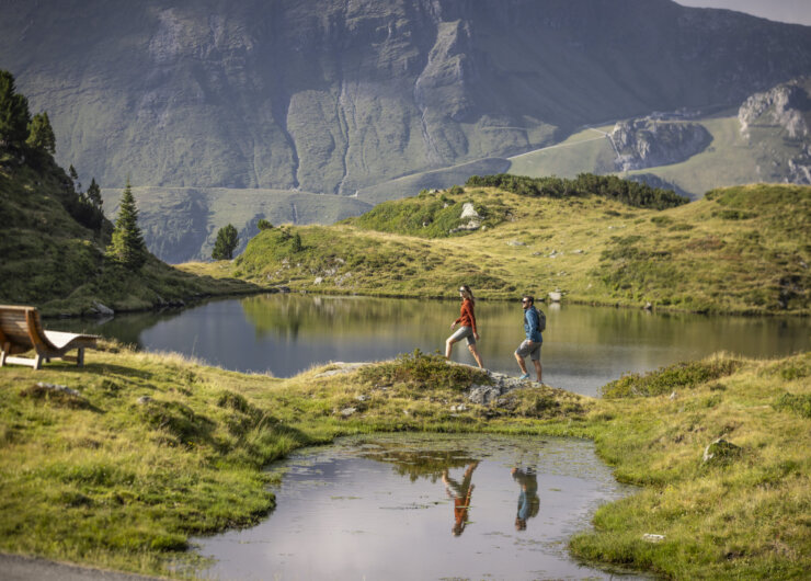 Twee mensen wandelen langs een klein bergmeer, omgeven door groene heuvels en bergen in de verte.