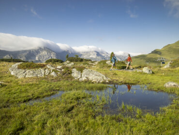 Twee wandelaars lopen over een grasachtig berglandschap met rotsen, een vijver en wolken die verre bergtoppen bedekken.