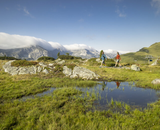Twee wandelaars lopen over een grasachtig berglandschap met rotsen, een vijver en wolken die verre bergtoppen bedekken.
