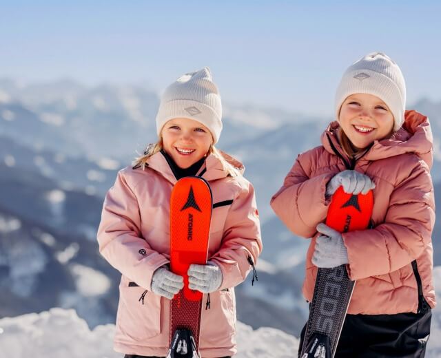 Dwoje uśmiechniętych dzieci w zimowych kurtkach z nartami, gotowych do zabawy z dziećmi w zaśnieżonych górach pod czystym niebem.