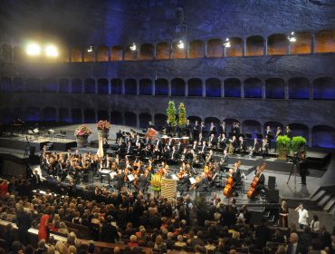 Orkiestra symfoniczna na scenie w wielkiej, kamiennej sali koncertowej z publicznością siedzącą z przodu.