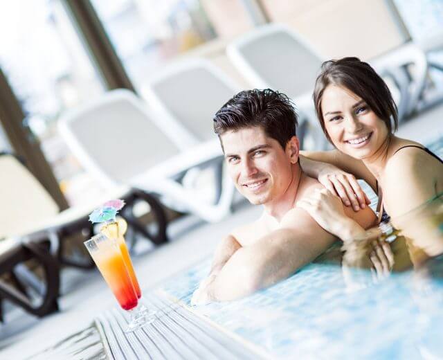 salzburger-thermen-wasserreiches-salzburgerland Uśmiechnięta para relaksująca się w basenie z kolorowym koktajlem na brzegu w pobliżu.