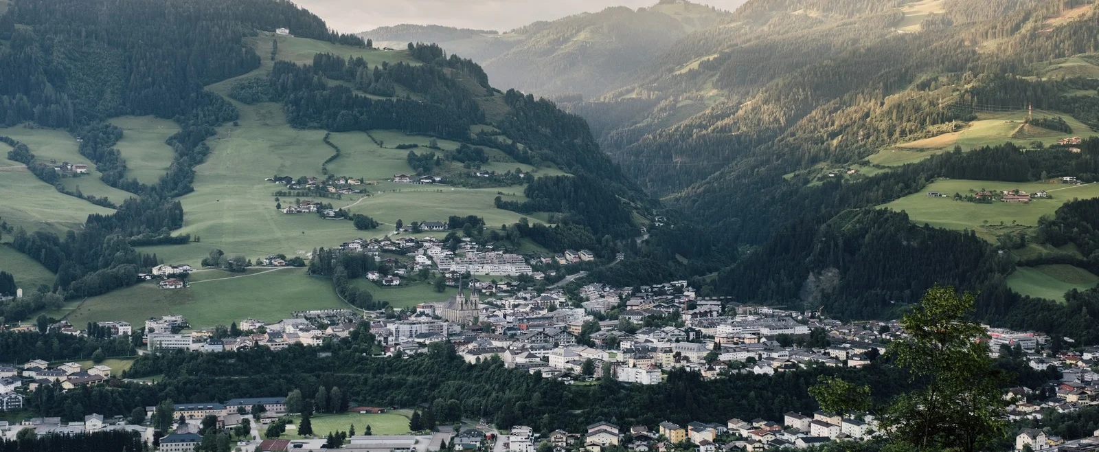 23_st_johann_in_salzburg_sommer_st_johann_ortsansicht_3-1600×658.jpg Małe miasteczko St. Johann na Ziemi Salzburskiej położone w zielonej dolinie pośród zalesionych gór i falistych wzgórz.