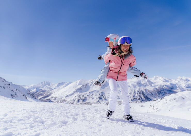 Kobieta daje dziecku przejażdżkę na barana podczas jazdy na nartach-Przegląd regionów na ośnieżonej górze pod czystym błękitnym niebem.