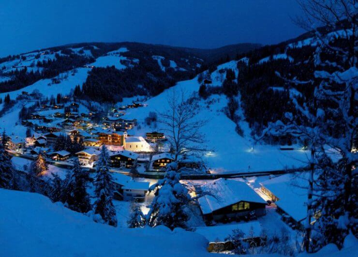 viehhofen-winter-panorama-nacht-1024×569 Pokryta śniegiem górska wioska o zmierzchu, z ciepłymi światłami świecącymi z domów i drzew otaczających okolicę.