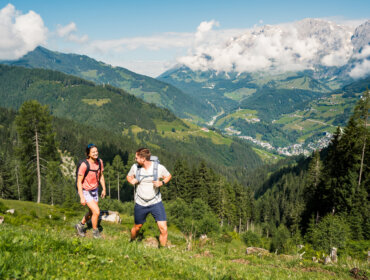 2024-07-11 Hochkönig Wander-Kampagne in Mühlbach HighRes-10 Dwóch wędrowców z plecakami idzie pod górę trawiastym szlakiem górskim Hochkönig, w tle malownicza dolina.