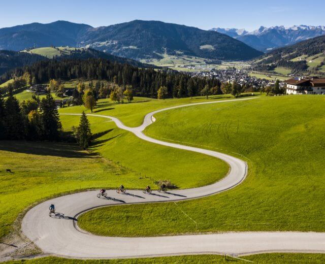 altenmarkt-zauchensee-bike-jonathan-forsthuber Rowerzyści jadą krętą drogą w Lato w Salzburskim Świecie Sportu, mijając zielone wzgórza, góry i wioskę.
