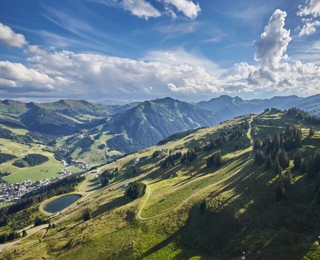 Widok z lotu ptaka na zielone góry z rozrzuconymi drzewami, małą wioskę i staw pod częściowo zachmurzonym niebem.